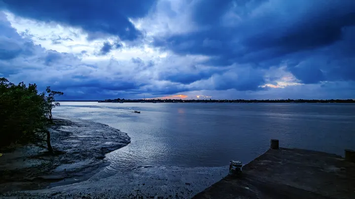 Wide Sundarbans estuary at blue hour with storm clouds, mudflats, and a distant boat silhouette.